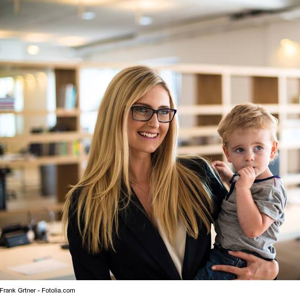 Eine blonde Frau in Business-Outfit trägt ihr kleines Kind im Arm durch die Büroumgebung.