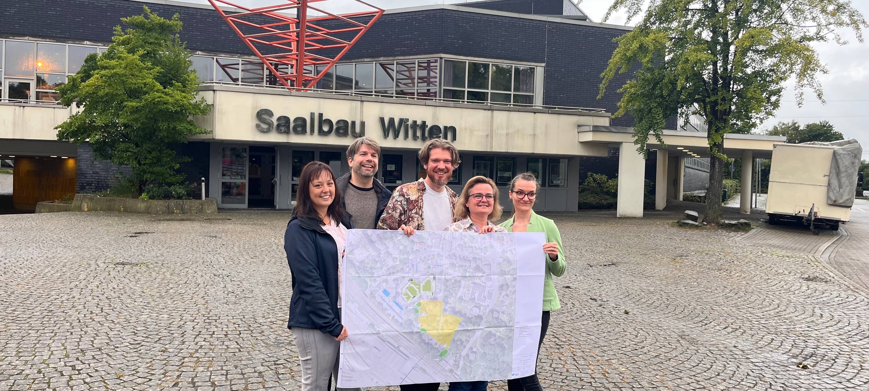 zu sehen sind 5 Personen, die vor dem Gebäude einen Bebauungsplan Plan halten.