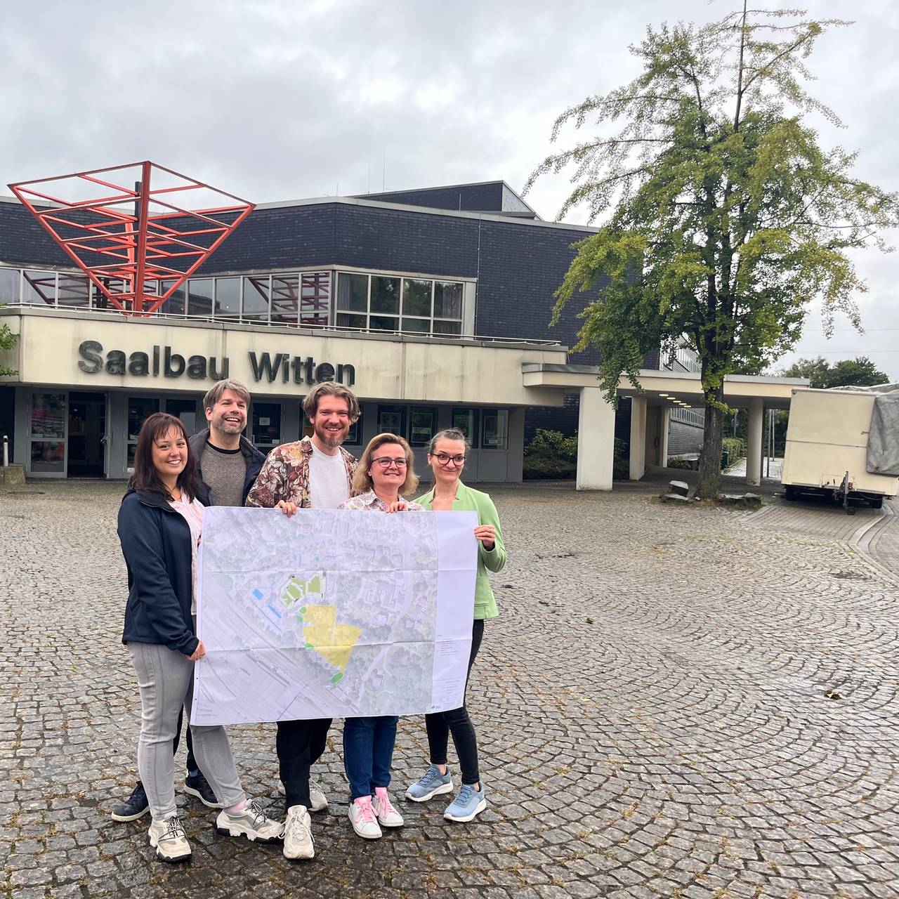 zu sehen sind 5 Personen, die vor dem Gebäude einen Bebauungsplan Plan halten.